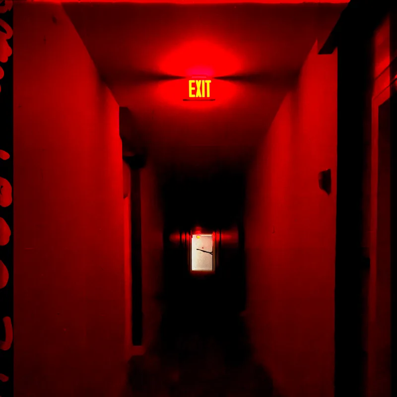 A dim corridor washed in deep red light, with a lit EXIT sign glowing above a dark doorway at the far end.